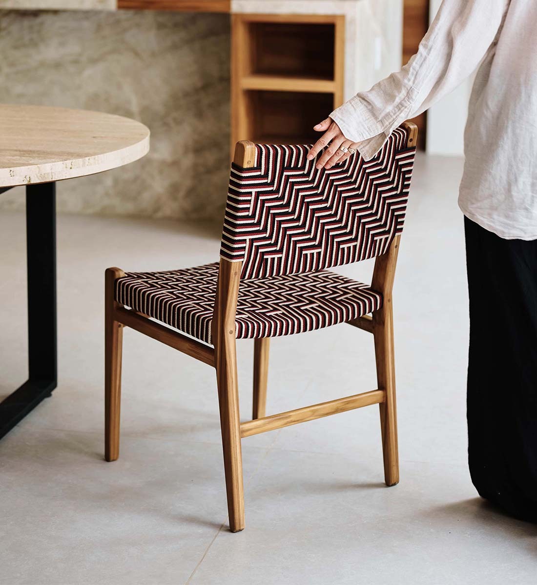 A mid-century modern wooden dining chair with a brown and white geometric woven seat and back, shown in a dining room setting with a person's hand resting on the backrest.